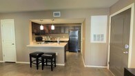 This single-floor space is designed with an open and functional layout that seamlessly connects the kitchen to the living area. The kitchen is a standout feature, showcasing rich dark wood cabinetry that extends along the walls, providing ample storage. A light-toned countertop contrasts the darker elements, creating a balanced aesthetic. The kitchen is equipped with black appliances, including a refrigerator and range, complemented by an integrated sink and pendant lighting that hangs gracefully above the peninsula. The breakfast bar, fitted with two dark cushioned stools, serves as both a casual dining spot and a natural divider between the kitchen and the adjacent living space. The floors throughout are finished in a smooth, neutral concrete-style surface that adds an understated, modern touch. Neutral warm-toned walls contribute to an inviting and cohesive ambiance throughout the space. A large wall vent and visible entryway hint at a practical, well-ventilated floor plan. Natural light filters in subtly, enhancing the overall warmth of the interior. The thoughtful design of this space prioritizes both comfort and utility, making it well-suited for everyday living.