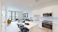 This thoughtfully designed single-floor residence showcases a clean, contemporary aesthetic with light hardwood flooring, crisp white walls, and floor-to-ceiling black-framed windows that flood the space with natural light and offer urban views. The open-concept kitchen is a standout feature, outfitted with white shaker cabinetry, a white subway tile backsplash, stainless steel appliances including a refrigerator, dishwasher, microwave, and range, and a generous white quartz island with bar seating. A sleek linear pendant light anchors the kitchen, which flows seamlessly into the living area. The living room is bright and welcoming, with large windows framing city rooftop views and ample space for comfortable seating arrangements. Two bedrooms continue the modern theme, each featuring floor-to-ceiling windows that bring in abundant daylight and neighborhood views, along with warm hardwood floors and clean wall finishes. The bathroom is elegantly appointed with large-format grey tile, a spacious walk-in glass-enclosed shower with built-in shelving, and polished fixtures with brushed gold accents. Throughout the residence, the design balances minimalist sensibility with warm, livable touches.