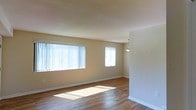 This single-floor space offers a clean, open layout highlighted by warm-toned hardwood flooring that extends throughout the main living area. The room is well-lit by multiple windows, including a large picture window dressed with vertical blinds that allows generous amounts of natural light to flood the interior, casting bright patches across the floor. A second, smaller window on the adjacent wall further enhances the airy feel of the space. The walls are finished in a neutral, warm beige tone that complements the natural wood flooring and contributes to an inviting ambiance. A pendant light fixture hangs near what appears to be a transitional area, suggesting a defined boundary between the living space and an adjoining room or dining area. White baseboards provide a crisp contrast to both the walls and the flooring. The overall design is straightforward and functional, with minimal architectural ornamentation, making the space highly adaptable for a variety of furniture arrangements and interior styles. The abundance of natural light and neutral palette create a bright, versatile environment.