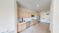 This single-floor unit showcases a clean, contemporary design throughout. The kitchen serves as a standout feature, offering an extended run of light maple cabinetry — both upper and lower — paired with white countertops and a white tile backsplash. Stainless steel appliances include a built-in microwave, dishwasher, and refrigerator, complemented by a black-finish sink faucet that adds a modern contrast. Recessed lighting illuminates the space efficiently. The open layout connects the kitchen to a generously sized living area, which benefits from large windows that frame outdoor views and allow natural light to fill the room. Light-toned vinyl flooring flows consistently throughout, contributing to the unit's cohesive, contemporary aesthetic. The bathroom is well-proportioned, featuring a combined tub and shower with built-in shelving, a toilet, and a single-sink vanity with light wood cabinetry, a white countertop, a matte black faucet, and a large mirror above. The overall design palette — white walls, light wood tones, and neutral flooring — creates a bright, airy ambiance that feels both functional and inviting.