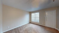 This single-floor unit is designed with a practical, open layout that connects the main living areas seamlessly. The living room is a well-proportioned space finished with wall-to-wall carpet in a neutral tan tone, complemented by warm beige walls and clean white baseboards. A ceiling fan provides overhead airflow, and a large window fitted with horizontal blinds allows natural light to fill the room. A white entry door adds a bright, welcoming touch to the space.

An adjacent carpeted room, visible through the floor plan, functions as a bedroom and also features a ceiling fan with an integrated light fixture, a window for natural light, and a closet with louvered doors. The textured ceilings are consistent throughout the unit.

The kitchen is accessible from the main living area and features tile flooring, white cabinetry, and white appliances including a range. A pass-through doorway connects the kitchen to the rest of the unit efficiently.

The bathroom is equipped with a full tub and shower combination surrounded by white tile, warm-toned floor tiles, a toilet, a vanity with storage, and wood-toned overhead cabinetry, providing functional and organized storage. The overall ambiance of the unit is clean and neutral.