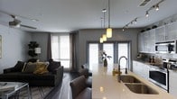 Located at 3001 Village Market Pl in Morrisville, NC, this single-floor residence offers a thoughtfully designed open-concept living space that blends modern aesthetics with everyday functionality. The living and kitchen areas flow seamlessly into one another, creating an expansive, connected feel. The kitchen is a standout feature, showcasing a large quartz-topped island with an undermount sink, a professional-style pull-down faucet, and crisp white cabinetry. A trio of pendant lights with warm-toned shades hangs elegantly above the island, complemented by track lighting that further illuminates the space. The living area is anchored by dark hardwood flooring and features a comfortable seating arrangement centered on a patterned area rug. Large windows dressed with layered curtains and horizontal blinds allow natural light to fill the room while offering privacy. The walls are finished in a contemporary cool gray tone throughout, lending the space a cohesive and sophisticated ambiance. Recessed ceiling lighting adds a clean, modern touch to the overall design. The open layout and thoughtful details make this a welcoming and versatile space suited for both relaxed living and entertaining.