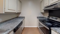 This single-floor space offers a well-organized layout across several distinct rooms, each finished with practical, low-maintenance materials. The living area features rich, dark wood-look flooring, neutral wall tones, and a ceiling fan with integrated lighting. A narrow window fitted with horizontal blinds allows natural light to filter in, while a white paneled entry door provides direct access to the unit.

Adjacent to the living area, a dedicated dining space is defined by warm wood-look flooring and an elegant brushed-nickel chandelier suspended from the ceiling, creating a focused area ideal for a dining table setup.

The galley-style kitchen is efficiently arranged with cream-toned cabinetry lining both sides, dark granite-look countertops, a built-in electric range, a dishwasher, and a ventilation hood above the cooking surface. Light wood flooring ties the kitchen together with a clean, functional aesthetic.

The bedrooms are carpeted for comfort and each features a ceiling fan with lighting and windows with blinds that bring in natural light. At least one bedroom includes a closet with built-in shelving for storage.

The bathroom includes wood-look flooring, a standard tub-shower combination, and clean, neutral walls throughout. White trim and baseboards unify the overall presentation of the space.