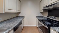 This single-floor space offers a well-organized layout across several distinct rooms, each finished with practical, low-maintenance materials. The living area features rich, dark wood-look flooring, neutral wall tones, and a ceiling fan with integrated lighting. A narrow window fitted with horizontal blinds allows natural light to filter in, while a white paneled entry door provides direct access to the unit.

Adjacent to the living area, a dedicated dining space is defined by warm wood-look flooring and an elegant brushed-nickel chandelier suspended from the ceiling, creating a focused area ideal for a dining table setup.

The galley-style kitchen is efficiently arranged with cream-toned cabinetry lining both sides, dark granite-look countertops, a built-in electric range, a dishwasher, and a ventilation hood above the cooking surface. Light wood flooring ties the kitchen together with a clean, functional aesthetic.

The bedrooms are carpeted for comfort and each features a ceiling fan with lighting and windows with blinds that bring in natural light. At least one bedroom includes a closet with built-in shelving for storage.

The bathroom includes wood-look flooring, a standard tub-shower combination, and clean, neutral walls throughout. White trim and baseboards unify the overall presentation of the space.