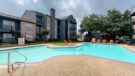 This residential community showcases a well-appointed outdoor amenity area centered around a generously sized swimming pool with clear turquoise water. The pool deck is finished in a textured aggregate surface, providing both durability and slip resistance, and is furnished with bright red lounge chairs positioned to take advantage of sunlight and open views. The surrounding landscape includes mature trees and maintained green lawns, adding a natural, shaded retreat to the outdoor space. The multi-story residential buildings framing the pool area feature a combination of dark teal and tan siding, pitched rooflines, and prominent chimney structures. Each building offers private balconies with railings, providing residents with personal outdoor space overlooking the courtyard. A wrought iron perimeter fence defines the pool area, enhancing both safety and aesthetic appeal. The overall ambiance is relaxed and community-oriented, with the layout encouraging outdoor gathering and leisure. The combination of mature landscaping, architectural variety, and resort-style pool amenities creates an inviting atmosphere suited for comfortable residential living.