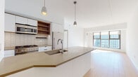 This single-floor residence showcases a contemporary design with clean lines and a neutral palette throughout. The open-plan kitchen and living area forms the heart of the space, where light hardwood flooring extends seamlessly underfoot, enhancing the sense of openness. The kitchen is anchored by a substantial island with a taupe quartz countertop, an integrated sink, and matte black hardware on white cabinetry. Stainless steel appliances—including a gas range, refrigerator, and microwave—are complemented by warm wood open shelving and a textured tile backsplash, adding visual depth. Pendant lighting above the island provides focused illumination, while the adjacent living area benefits from generous floor-to-ceiling windows that invite abundant natural light and offer expansive urban views. The bathroom is thoughtfully finished with contrasting hexagonal tiles—white on the walls and dark on the floor—paired with a freestanding soaking tub, a floating vanity with wood accents, and a large mirror cabinet. A dedicated in-unit laundry closet with louvered doors provides practical convenience. Throughout, the space maintains a cohesive modern aesthetic with high ceilings and a bright, airy ambiance.