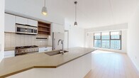 This single-floor residence showcases a contemporary design with clean lines and a neutral palette throughout. The open-plan kitchen and living area forms the heart of the space, where light hardwood flooring extends seamlessly underfoot, enhancing the sense of openness. The kitchen is anchored by a substantial island with a taupe quartz countertop, an integrated sink, and matte black hardware on white cabinetry. Stainless steel appliances—including a gas range, refrigerator, and microwave—are complemented by warm wood open shelving and a textured tile backsplash, adding visual depth. Pendant lighting above the island provides focused illumination, while the adjacent living area benefits from generous floor-to-ceiling windows that invite abundant natural light and offer expansive urban views. The bathroom is thoughtfully finished with contrasting hexagonal tiles—white on the walls and dark on the floor—paired with a freestanding soaking tub, a floating vanity with wood accents, and a large mirror cabinet. A dedicated in-unit laundry closet with louvered doors provides practical convenience. Throughout, the space maintains a cohesive modern aesthetic with high ceilings and a bright, airy ambiance.