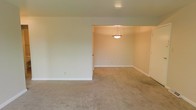 This single-story space presents a straightforward, functional layout characterized by neutral design choices throughout. The interior showcases wall-to-wall beige carpeting that extends across both the main living area and the adjoining room, creating a cohesive and comfortable feel underfoot. Walls are finished in a clean, neutral off-white tone, contributing to a bright and airy atmosphere.

The layout features an open connection between two distinct areas — a spacious living zone and an adjoining room, likely used as a dining area, separated by a wide pass-through opening rather than a full wall. This design choice enhances the sense of openness and allows light to flow freely between the two spaces.

A pendant light fixture is suspended from the ceiling in the adjoining room, providing a subtle focal point and functional overhead lighting. Recessed lighting in the main area supplements the ambient light. Clean white baseboards frame the perimeter of each room, adding a crisp finishing detail. The overall condition appears well-maintained, presenting a blank canvas ready for personalization.