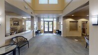Located at 4701 Seventh Ave SW in Olympia, Washington, this senior living community is designed to offer comfort, community, and a rich array of amenities all on a single level. The grand entrance lobby sets an impressive tone with soaring double-height ceilings, clerestory windows that draw in natural light, and elegant wall sconces flanking framed artwork. Seating areas near the reception desk provide a welcoming atmosphere for residents and visitors alike. Dual water fountains are conveniently positioned along interior corridors. The library and lounge area is a cozy retreat, featuring built-in bookshelves stocked with a wide selection of titles, a fireplace, comfortable sofas, and views of the lush outdoor grounds. A spacious dining room is anchored by structural columns and accented with a fireplace, offering residents an inviting setting for meals at well-appointed tables. A casual bistro-style lounge provides additional seating with vibrant red upholstered chairs, booth seating, and ample natural light. For entertainment, a dedicated private theater room features tiered rows of plush leather recliner-style seating, classic movie poster décor, and a projection screen. Long, well-lit corridors connect the various wings of the community, ensuring easy navigation throughout the building.
