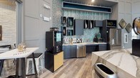 This thoughtfully designed clubhouse delivers a welcoming community gathering space that blends upscale aesthetics with everyday functionality. The single-floor layout encompasses several distinct zones that flow seamlessly together, unified by wood-look plank flooring and a cohesive contemporary design palette.

The main lounge area commands attention with soaring ceilings and dramatic floor-to-ceiling windows that flood the space with natural light and frame views of the outdoor pool and courtyard. Plush sectional sofas and leather club chairs are arranged around dark wood coffee tables, while wainscoted walls in soft grey provide an elegant backdrop for curated original artwork.

A dedicated media lounge offers a wall-mounted flat-screen television flanked by teal accent shelving, creating an intimate entertainment nook. Adjacent dining and bar-height seating areas provide flexible spaces for casual gatherings, all bathed in natural light from expansive poolside windows.

The kitchenette is a standout feature, showcasing a striking teal mosaic tile backsplash, dark cabinetry, marble-look countertops, a stainless steel dishwasher, and a dedicated Starbucks coffee machine. A textured white stacked-stone accent wall adds warmth and dimension throughout the common areas, complemented by rustic wood décor elements and curated barware-themed wall art, creating an overall ambiance that is both refined and welcoming.