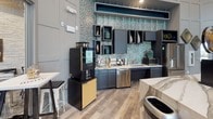 This thoughtfully designed clubhouse delivers a welcoming community gathering space that blends upscale aesthetics with everyday functionality. The single-floor layout encompasses several distinct zones that flow seamlessly together, unified by wood-look plank flooring and a cohesive contemporary design palette.

The main lounge area commands attention with soaring ceilings and dramatic floor-to-ceiling windows that flood the space with natural light and frame views of the outdoor pool and courtyard. Plush sectional sofas and leather club chairs are arranged around dark wood coffee tables, while wainscoted walls in soft grey provide an elegant backdrop for curated original artwork.

A dedicated media lounge offers a wall-mounted flat-screen television flanked by teal accent shelving, creating an intimate entertainment nook. Adjacent dining and bar-height seating areas provide flexible spaces for casual gatherings, all bathed in natural light from expansive poolside windows.

The kitchenette is a standout feature, showcasing a striking teal mosaic tile backsplash, dark cabinetry, marble-look countertops, a stainless steel dishwasher, and a dedicated Starbucks coffee machine. A textured white stacked-stone accent wall adds warmth and dimension throughout the common areas, complemented by rustic wood décor elements and curated barware-themed wall art, creating an overall ambiance that is both refined and welcoming.