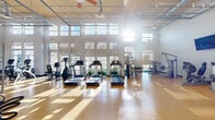 Located at 900 Grange Hall Dr in Euless, TX, this fitness center offers a generously sized, single-floor exercise space designed to accommodate a wide range of workout routines. The facility features soaring ceilings with a suspended grid system and large ceiling fans that promote airflow throughout the open floor plan. Walls of tall, multi-pane windows flood the interior with abundant natural light, creating a bright and energizing atmosphere while offering views of the surrounding outdoors.

The exercise room is outfitted with a comprehensive selection of equipment, including multiple treadmills, stationary bikes, and elliptical machines arranged along the windowed wall for a motivating workout environment. A dedicated strength training area includes Hoist-branded selectorized machines targeting various muscle groups, a functional training rig with battle ropes, medicine balls, kettlebells, and a heavy bag. Wall-mounted flat-screen televisions are positioned throughout the space for entertainment during workouts.

The flooring throughout consists of warm-toned hardwood-style material that is both durable and visually appealing. Full-length mirrors in the strength area enhance the sense of space and support proper form. The facility is accessible via glass double doors, and the open layout ensures easy navigation between cardio, strength, and functional training zones.