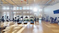 Located at 900 Grange Hall Dr in Euless, TX, this fitness center offers a generously sized, single-floor exercise space designed to accommodate a wide range of workout routines. The facility features soaring ceilings with a suspended grid system and large ceiling fans that promote airflow throughout the open floor plan. Walls of tall, multi-pane windows flood the interior with abundant natural light, creating a bright and energizing atmosphere while offering views of the surrounding outdoors.

The exercise room is outfitted with a comprehensive selection of equipment, including multiple treadmills, stationary bikes, and elliptical machines arranged along the windowed wall for a motivating workout environment. A dedicated strength training area includes Hoist-branded selectorized machines targeting various muscle groups, a functional training rig with battle ropes, medicine balls, kettlebells, and a heavy bag. Wall-mounted flat-screen televisions are positioned throughout the space for entertainment during workouts.

The flooring throughout consists of warm-toned hardwood-style material that is both durable and visually appealing. Full-length mirrors in the strength area enhance the sense of space and support proper form. The facility is accessible via glass double doors, and the open layout ensures easy navigation between cardio, strength, and functional training zones.