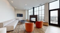 This well-appointed communal lounge space spans a generous single floor, thoughtfully designed to serve as a sophisticated gathering and relaxation area. The layout is divided into two distinct functional zones: a lounge area and a kitchenette, connected by an open, airy floor plan.

The lounge area is the visual centerpiece of the space, anchored by a striking double-sided fireplace encased in a dark metal surround with a cylindrical flue extending to the ceiling. The fireplace is positioned against a dramatic wall of floor-to-ceiling steel-framed windows, which flood the interior with natural light and offer views of an outdoor terrace. Built-in wood bench seating lines the window wall, complementing the textured, multi-toned carpet underfoot. Furnishings include a long, low-profile light gray sofa, round matte charcoal side tables, and bold terracotta-orange swivel chairs that add a warm contrast. A wall-mounted flat-screen television completes the media setup.

The kitchenette features light hardwood flooring, matte gray cabinetry with black hardware, a dark stone countertop, a matte black faucet, a stainless steel sink, a dishwasher, and a warming drawer. A glossy white subway tile backsplash and floating wood shelves add a polished, modern finish. Recessed lighting throughout ensures consistent, even illumination across both zones.