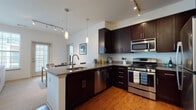 This well-designed single-floor residence offers a cohesive blend of modern finishes and functional living spaces. The open-concept kitchen is a standout feature, showcasing rich dark espresso cabinetry, granite countertops, and a full suite of stainless steel appliances including a gas range, built-in microwave, and dishwasher. A kitchen island with an integrated sink provides additional prep space and doubles as a casual dining area, while pendant lighting and track lighting illuminate the space with a warm, inviting glow. Warm hardwood flooring flows throughout the kitchen and into the adjacent living area, which is bathed in natural light from a generous bank of large windows. The living space offers a relaxed, neutral palette ideal for comfortable everyday living. The residence includes two bedrooms, each carpeted and filled with natural light from dual windows. One bedroom features a tufted upholstered headboard and elegant dual bedside lamps, lending a refined feel. The bathrooms are finished with dark vanity cabinetry, light tile flooring, and white countertops, with one bathroom offering a shower-tub combination. A well-placed half bath serves as a convenient guest space. Throughout, the design maintains a consistent, contemporary aesthetic.