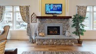 This single-floor common area blends farmhouse charm with modern comfort across a generously proportioned open layout. Light wood-tone flooring runs throughout, unifying the various functional zones seamlessly. The main lounge area is anchored by a striking floor-to-ceiling stone fireplace with a rustic wood mantel and a mounted television above, creating a warm and inviting focal point. Vaulted ceilings add a sense of openness and airiness to the space.

A seating nook near the large, multi-pane windows features elegant wingback chairs with nailhead trim, a round bistro table, and an industrial-style adjustable stool, all bathed in generous natural light and views of the surrounding greenery and neighboring buildings. Patterned curtains and wall art contribute a layered, curated aesthetic.

The kitchenette area is finished with dark cabinetry, light stone-look countertops, a stainless steel sink, dishwasher, and a single-serve coffee maker, all accented by farmhouse-themed wall décor. The central lounge accommodates plush leather seating arranged around a coffee table, making the space well-suited for relaxed gatherings and social interaction.