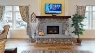 This single-floor common area blends farmhouse charm with modern comfort across a generously proportioned open layout. Light wood-tone flooring runs throughout, unifying the various functional zones seamlessly. The main lounge area is anchored by a striking floor-to-ceiling stone fireplace with a rustic wood mantel and a mounted television above, creating a warm and inviting focal point. Vaulted ceilings add a sense of openness and airiness to the space.

A seating nook near the large, multi-pane windows features elegant wingback chairs with nailhead trim, a round bistro table, and an industrial-style adjustable stool, all bathed in generous natural light and views of the surrounding greenery and neighboring buildings. Patterned curtains and wall art contribute a layered, curated aesthetic.

The kitchenette area is finished with dark cabinetry, light stone-look countertops, a stainless steel sink, dishwasher, and a single-serve coffee maker, all accented by farmhouse-themed wall décor. The central lounge accommodates plush leather seating arranged around a coffee table, making the space well-suited for relaxed gatherings and social interaction.