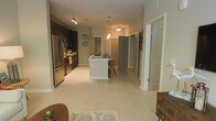 This single-floor space is designed with an open-concept layout that seamlessly connects the kitchen and living areas, creating a spacious and functional environment. The kitchen is a standout feature, equipped with stainless steel appliances, including a full-size refrigerator, and dark cabinetry that provides a striking contrast against the light-toned tile flooring that extends throughout the unit. A central white kitchen island serves as the focal point of the space, offering additional counter space and seating with bar-style chairs positioned along its side. Pendant lighting hangs above the island, complementing the track lighting installed over the main kitchen area, both contributing to a warm, well-lit ambiance. The neutral-toned walls and light tile flooring enhance the sense of openness and brightness. Toward the rear of the space, multiple doorways lead to additional rooms, suggesting a practical and well-organized floor plan. Small decorative touches, including framed artwork on the walls and patterned area rugs, add personality to the otherwise clean and contemporary aesthetic. Overall, the space balances modern design with everyday functionality.