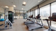 This contemporary fitness center is designed to deliver a comprehensive workout experience in a bright, open environment. The space is defined by expansive floor-to-ceiling windows that line the perimeter, flooding the interior with natural light and offering sweeping views of the surrounding cityscape and greenery — an energizing backdrop for any workout session.

The cardio zone is well-appointed with a row of treadmills, elliptical trainers, and stationary bikes, all featuring individual screens and positioned to take full advantage of the outdoor views. The strength training area is equally comprehensive, offering a selection of Precor cable machines, functional trainers, weight benches, and a dedicated free weights section stocked with a full rack of dumbbells and stability balls for functional training.

A glass-walled corridor and elegant pendant light installation connect the fitness area to an adjacent space, adding an architectural flair that elevates the overall aesthetic. Striped accent walls introduce a pop of color and visual interest, complementing the otherwise neutral, modern palette of the room.

The space features a drop tile ceiling with recessed and panel lighting throughout, ensuring consistent illumination across all workout zones. Plush carpet flooring in the main training areas provides comfort underfoot, while the overall layout allows for ample movement and spacing between equipment, creating a welcoming and functional fitness environment.