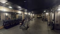 Located at 1515 W Monroe St in Chicago's West Loop neighborhood, this fitness facility occupies a single expansive floor designed to accommodate a comprehensive range of workout disciplines. The space draws on an industrial aesthetic, with exposed brick walls, dark painted ceilings, and polished concrete flooring that lend a moody, club-like atmosphere throughout.

The cardio zone lines one side of the facility with a row of treadmills, stair climbers, and other cardio machines, each equipped with personal screens. Adjacent to this area, a long turf lane flanked by wall-mounted functional training equipment and medicine ball racks provides ample space for athletic conditioning and functional movement exercises.

The free weights area is anchored by extensive dumbbell racks, multiple adjustable benches, barbell stations, and a squat rack. Full-length mirrors along the walls expand the sense of space and allow for form monitoring. Rubber flooring in this zone provides both durability and safety.

A barre studio area features floor-to-ceiling mirrors and freestanding ballet barres, reflecting the broader facility and adding a sense of depth. A separate lounge area offers a distinct retreat, furnished with a black leather sectional, leather accent chair, botanical-patterned wallpaper, lush plants, and framed artwork, creating a relaxed recovery environment.