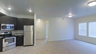 This single-floor unit offers an open-concept design that seamlessly connects the kitchen and living areas, creating a bright and functional living space. The kitchen is equipped with dark cabinetry that provides a striking contrast against the light-toned walls, and features stainless steel appliances, including a full-size refrigerator. Stone-finish countertops add a modern touch, while a structural column subtly defines the boundary between the kitchen and the main living area.

The living area is generously sized, offering ample room for a variety of furniture arrangements. Light hardwood-style flooring runs throughout, contributing to a clean, cohesive aesthetic. Recessed and flush-mount ceiling lighting illuminate the space evenly, enhancing the airy feel of the open floor plan.

A hallway visible from the main area leads to additional rooms, suggesting a practical layout with clear separation between living and private spaces. White interior doors and crisp white walls complement the neutral palette, giving the unit a fresh, move-in-ready appearance. Overall, the space balances modern finishes with a simple, welcoming ambiance.