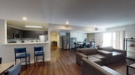 This single-floor residence offers a well-organized, functional layout designed to accommodate multiple occupants comfortably. The open-concept common area seamlessly connects the living room, dining space, and kitchen, creating a communal hub ideal for shared living. The living room features grey upholstered sofas, a coffee table, and shelving units set on warm hardwood-style flooring beneath recessed lighting. The dining area provides ample seating with multiple tables and chairs, and is brightened by a large window with natural light. The fully equipped kitchen includes stainless steel appliances—a refrigerator, electric range, and microwave—paired with dark cabinetry, a tiled backsplash, and a breakfast bar with bar seating. Multiple bedrooms are furnished with twin beds dressed in blue bedding, individual desks, and chairs to support both rest and study. Each bedroom offers a clean, efficient layout with ample space for personal use, and some feature direct access to additional adjoining rooms. The bathrooms maintain a modern aesthetic with dark wood vanities, vessel sinks, quartz-style countertops, large mirrors, and wood-look flooring, providing a polished and practical finish throughout the unit.