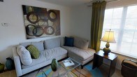 This single-floor space centers around a thoughtfully designed living area that balances comfort and style. The room features a spacious sectional sofa in a soft neutral gray, adorned with accent pillows in deep teal and textured green, adding pops of color against the clean, light-colored walls. A large, bold piece of abstract wall art anchors the space, showcasing overlapping circular motifs in rich earth tones that draw the eye and set a contemporary tone. Natural light filters into the room through a window dressed with floor-length olive-green drapery, complemented by a set of horizontal blinds that allow for adjustable light control. A glass-top coffee table with a chrome frame sits at the center of the seating area, topped with a decorative book and a sculptural dark green vase, adding an artistic touch. A warm-toned table lamp on a rich wood side table provides ambient lighting, enhancing the cozy atmosphere. The flooring features a multi-toned area rug in blues, greens, and yellows that ties the color palette together. Overall, the space exudes a relaxed yet curated aesthetic, with intentional design choices that make it feel both livable and visually engaging.