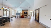 Located at 1824 Defoor Ave NW in Atlanta, Georgia, this single-floor community clubhouse offers a generously sized, open-plan layout designed for comfort and social interaction. The space is anchored by soaring vaulted ceilings that amplify the sense of openness, while wide-plank wood-look tile flooring flows seamlessly throughout, lending warmth and continuity to the design.

At the heart of the space, a dramatic stone accent wall frames a wall-mounted TV and a stacked-wood fireplace, creating a striking focal point for the lounge area, which features plush neutral seating arranged for conversation. Adjacent to the lounge, a sleek dark-toned bar counter with bar stools sits beneath a trio of stylish pendant lights, complemented by large windows that draw in abundant natural light and views of the surrounding greenery.

A separate seating alcove with swivel armchairs and a round side table offers a more intimate retreat, while a mirrored interior window connects visual space between areas, enhancing the open feel. A hallway with an ornate wrought-iron stair railing and a sliding barn door adds refined transitional detail. Recessed lighting and large framed windows throughout ensure the space remains bright and welcoming at all times.
