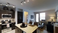 This single-floor residence showcases a thoughtfully designed open-concept layout that connects the kitchen and living areas into one cohesive space. The kitchen is anchored by a large granite-topped island with a light, veined finish, offering both meal preparation space and casual dining with seating for multiple guests. Dark, matte cabinetry lines the kitchen walls, providing a bold contrast against the light granite surfaces and pale grey walls. A tall, dark pantry cabinet adds significant storage presence to the space.

Track lighting along the kitchen ceiling delivers focused illumination throughout the cooking and dining zones, enhancing functionality and visual appeal. The open layout transitions naturally into the living area, which benefits from a large window that allows abundant natural light to fill the room. The living space comfortably accommodates seating and a coffee table, creating a relaxed yet polished atmosphere. A hallway visible from the main area hints at additional rooms beyond, including what appears to be a bathroom. Light hardwood-style flooring runs throughout, tying the kitchen and living zones together cohesively and contributing to the space's modern, clean aesthetic.