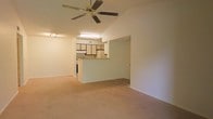 This single-floor space features an open-concept layout that connects the living area to the kitchen, creating a cohesive and functional floor plan. The living area is highlighted by vaulted ceilings that add a sense of height and openness, complemented by a ceiling fan with a light fixture that provides both air circulation and ambient lighting. A track lighting fixture is also present along the upper wall, offering additional illumination options.

The flooring throughout the living area consists of neutral-toned carpeting, contributing to a warm and understated aesthetic. The walls are finished in a light, neutral tone that enhances the brightness of the space.

The kitchen is positioned at the rear of the living area and is partially separated by a raised pass-through bar or peninsula, which provides a natural visual division while maintaining the open feel. The kitchen features white cabinetry with contrasting darker accent panels on the upper cabinets, a skylight or overhead fluorescent fixture for added brightness, and standard appliances including a refrigerator. The overall ambiance of the space is straightforward and functional, with a clean, move-in-ready presentation.