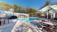 Located at 500 Palisades Drive in Birmingham, Alabama, this outdoor pool area is a well-appointed communal retreat designed for relaxation and leisure. The centerpiece is a generously sized freeform swimming pool with crystal-clear turquoise water, accessible via stainless steel entry rails and surrounded by a broad, light-toned tiled pool deck that provides ample space for lounging.

The area is framed by a series of classic white pergolas that line the perimeter, offering shaded seating areas furnished with lounge chairs and umbrella-shaded tables — ideal for outdoor dining or unwinding poolside. A prominent stone retaining wall runs along one side, adding a refined, architectural character to the space and integrating natural textures into the overall design.

The backdrop of mature trees and lush greenery creates a private, serene atmosphere, while the fully fenced enclosure ensures security and seclusion. Adjacent to the pool stands an elegant two-story clubhouse building with brick and siding exteriors, a covered upper balcony, and large windows that overlook the pool area, adding a stately architectural presence.

Under wide-open blue skies, this outdoor space strikes a balance between polished design and natural surroundings, making it an inviting venue for recreation and relaxation throughout the warmer months.