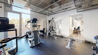 Located at 1 N Fifth Ave in downtown Tucson, Arizona, this fitness center is a well-equipped exercise room designed to support a full range of workout routines. The space features an industrial-style aesthetic, with exposed mesh ceilings, painted concrete block walls, and durable rubber flooring throughout — all contributing to a functional, no-frills gym atmosphere.

The room is outfitted with an impressive array of equipment for its size, including a treadmill, elliptical trainer, stationary spin bike, and a multi-station cable strength machine. A dedicated dumbbell rack with Vectra-branded free weights provides options for resistance training, while a wall-mounted pull-up bar adds a bodyweight training element. A weight bench and additional free weights round out the strength training offerings.

Large windows along one wall allow natural light to flood the space and offer views of the surrounding Tucson cityscape, creating an energizing workout environment. A wall-mounted flat-screen television provides entertainment during exercise sessions. Practical amenities include a water fountain and a paper towel dispenser, ensuring convenience for users. A full-length mirror enhances the sense of space while allowing users to monitor their form. Track lighting overhead ensures the room remains bright and well-illuminated throughout the day.