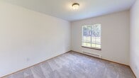 Located in Poulsbo, WA, this single-floor, two-bedroom, one-bathroom unit offers a practical and well-organized layout. The living room serves as the central gathering space, featuring light wood-tone laminate flooring, a ceiling fan, and a pass-through opening that connects directly to the kitchen, promoting an open feel despite the compact footprint. Large windows allow natural light to fill the living area, creating a bright and welcoming atmosphere.

The kitchen is efficiently designed with white cabinetry accented by wood-tone trim, gray countertops, a coil-burner electric range, and a refrigerator. Ample upper and lower cabinet storage ensures functional use of the available space.

The primary bedroom features a wide picture window with outdoor views of the surrounding greenery and a baseboard heater. The second bedroom is carpeted, includes a window, and is equipped with a sliding wood-panel closet offering organized storage.

The bathroom includes a vanity with gray countertop, a well-lit mirror, a medicine cabinet, and an adjacent open shelving unit providing additional storage. A central hallway connects all rooms, finished with the same hardwood-style flooring seen throughout the main living areas.