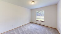 Located in Poulsbo, WA, this single-floor, two-bedroom, one-bathroom unit offers a practical and well-organized layout. The living room serves as the central gathering space, featuring light wood-tone laminate flooring, a ceiling fan, and a pass-through opening that connects directly to the kitchen, promoting an open feel despite the compact footprint. Large windows allow natural light to fill the living area, creating a bright and welcoming atmosphere.

The kitchen is efficiently designed with white cabinetry accented by wood-tone trim, gray countertops, a coil-burner electric range, and a refrigerator. Ample upper and lower cabinet storage ensures functional use of the available space.

The primary bedroom features a wide picture window with outdoor views of the surrounding greenery and a baseboard heater. The second bedroom is carpeted, includes a window, and is equipped with a sliding wood-panel closet offering organized storage.

The bathroom includes a vanity with gray countertop, a well-lit mirror, a medicine cabinet, and an adjacent open shelving unit providing additional storage. A central hallway connects all rooms, finished with the same hardwood-style flooring seen throughout the main living areas.