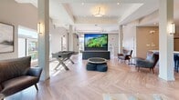 Located in Linley Valley, Nanaimo, BC, this single-floor amenity space offers a thoughtfully designed open-plan layout that seamlessly integrates a fully equipped kitchen with a spacious living area. The architecture is defined by soaring ceilings that amplify the sense of openness throughout, while expansive floor-to-ceiling windows flood the interior with abundant natural light and frame lush, tree-lined views of the surrounding landscape.

The kitchen is outfitted with contemporary concrete-finish cabinetry, stainless steel appliances including a gas range, dishwasher, and refrigerator, complemented by clean white countertops and a crisp white glass backsplash. Pendant lighting adds a refined finishing touch above the kitchen island. The herringbone-patterned hardwood flooring flows consistently throughout the space, adding warmth and visual texture.

The living area is generously proportioned and versatile, accommodating multiple seating arrangements including leather lounge chairs, sofas, and ottomans. A large projection screen serves as a focal point for entertainment, while a foosball table adds a recreational element. Curated artwork adorns the walls, enhancing the upscale ambiance.

An adjoining covered outdoor patio, finished with white vertical board cladding and recessed lighting, provides a comfortable extension of the interior living space, offering additional seating with views to the surrounding neighbourhood.