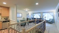 This two-story residence showcases a thoughtfully designed layout that balances both comfort and elegance. The main living level is anchored by an open-concept floor plan that seamlessly connects the dining area and living room, creating a spacious and cohesive environment. A wrought-iron staircase with decorative balusters and a light wood handrail serves as a striking architectural centerpiece, offering a view across the entire living space from the upper landing.

The living room is generously proportioned and filled with natural light streaming through large windows dressed with sheer, floor-length curtains in soft teal tones. The space accommodates a substantial seating arrangement, with dark upholstered sofas accented by blue throw pillows, complementing the cool, neutral color palette throughout. A glass-topped coffee table adds a refined touch to the lounge area.

Adjacent to the living room, the dining area features a round glass table surrounded by teal upholstered chairs, creating an intimate yet stylish setting for gatherings. A decorative framed mirror on the wall enhances the sense of depth and light in the space. Recessed and flush-mount ceiling fixtures provide warm, even illumination throughout. The overall ambiance is polished, airy, and welcoming, with a consistent design language that ties both floors together cohesively.