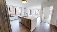 Located at 66 Summer St in downtown Stamford, Connecticut, this modern residence offers a thoughtfully designed single-floor layout with a clean, contemporary aesthetic. The open-concept kitchen serves as the centerpiece of the living area, anchored by a large island with a quartz countertop, an integrated sink with a gold-toned faucet, and wood-grain cabinetry that adds warmth to the space. Stainless steel appliances — including a range, over-the-range microwave, dishwasher, and refrigerator — complement the white subway tile backsplash and crisp white upper cabinets, creating a refined, functional cooking environment. The open living and dining area flows seamlessly from the kitchen, featuring hardwood-style flooring and oversized windows that flood the space with natural light and frame views of the surrounding urban streetscape. A private bedroom is carpeted for comfort and includes a generously sized walk-in closet with built-in shelving and hanging rods. The bathroom features large-format tile flooring, a floating wood-grain vanity with a white countertop, a framed mirror, and a wall-mounted light fixture, delivering a sleek, spa-inspired feel. Throughout the residence, the consistent use of neutral tones, modern finishes, and natural light contributes to a bright and inviting atmosphere.