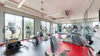 This fitness center at 2981 Olentangy River Road in Columbus, Ohio offers a thoughtfully designed exercise space across a single floor. The main exercise room is the focal point of the facility, featuring a striking red and gray rubberized floor that adds a bold, energetic aesthetic to the space. Large floor-to-ceiling windows line the walls, flooding the room with natural light and providing open views of the surrounding Columbus cityscape and greenery, including views toward neighboring buildings and tree-lined streetscapes. The high ceilings, accented by exposed HVAC ductwork, lend an industrial yet functional character to the space. Full-length mirrored walls expand the sense of openness and allow users to monitor their form during workouts. The room is well-equipped with a comprehensive array of cardio machines, including treadmills, ellipticals, stationary bikes, and rowing machines, all arranged to maximize floor space and traffic flow. Wall-mounted flat-screen televisions are positioned throughout for entertainment during workouts. Overhead linear lighting supplements the abundant natural light. A hallway connects to additional utility areas, and a water fountain station is conveniently available for hydration. The overall ambiance is modern, clean, and motivating.