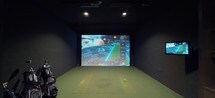 This single-floor entertainment space is divided into two distinct yet complementary areas: a professional-grade golf simulator bay and a refined lounge. The golf simulator bay is finished with dark walls and turf flooring, creating an immersive environment centered around a large projection screen running an advanced simulation system. Recessed ceiling lighting illuminates the hitting area, and a secondary display monitor is mounted to the side for additional game data. A golf bag station is positioned conveniently within the bay.

Adjacent to the simulator is a well-appointed lounge area that exudes a moody, sophisticated ambiance. Dark walls are paired with warm wood flooring in a parquet-style layout, and exposed ceiling beams are strung with Edison bulb lighting, adding an industrial yet inviting character. Seating includes a rich burgundy tufted leather Chesterfield sofa flanked by contemporary grey armchairs, arranged around an aviator-style metal coffee table atop a vintage patchwork rug. Framed artwork decorates the walls, and a built-in shelving unit with cabinetry and a low credenza provides practical storage, completing this stylish entertainment destination.