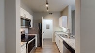 Located at 7101–7169 Libby Rd in Plano, TX, this single-floor residence offers a thoughtfully designed layout with functional living spaces throughout. The galley-style kitchen is a standout feature, showcasing white shaker cabinetry with brushed nickel hardware, granite-look countertops, and a full suite of stainless steel appliances including a smooth-top range, over-the-range microwave, refrigerator, and dishwasher. Wood-look flooring runs the length of the kitchen, complemented by track lighting overhead that provides ample task illumination. Adjacent to the kitchen, the living area transitions to soft carpeting, offering a comfortable and warm atmosphere. The space is kept neutral throughout with warm greige wall tones that provide a versatile backdrop. The bedroom areas are generously sized, each featuring plush carpet and windows dressed with horizontal blinds that allow for natural light while maintaining privacy. Views of surrounding greenery are visible from the bedroom windows. The bathroom includes a soaking tub and shower combination with distinctive green and black checkered tile accent detailing along the upper wall, adding a classic touch. The overall layout, as visible in the dollhouse view, is efficiently organized, with clearly defined zones for living, sleeping, and bathing.