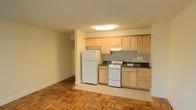 This single-floor space presents a functional and straightforward layout, with the kitchen serving as a central feature. The kitchen is equipped with light maple-toned wood cabinetry offering ample storage across both upper and lower units. Dark countertops provide a subtle contrast to the warm cabinetry, while a gray tile backsplash adds a clean, textured detail to the cooking area. White appliances, including a full-size refrigerator, a gas range, and an under-cabinet range hood, complete the setup. A stainless steel sink is positioned along the counter with easy access. The kitchen floor is finished in neutral tile, which transitions into the adjoining living area where classic parquet hardwood flooring adds warmth and character to the space. The open layout between the kitchen and living area creates a sense of connected, usable space. Flush-mount ceiling lighting illuminates both zones evenly. The walls are finished in a soft, neutral tone, contributing to a bright and understated overall ambiance throughout the unit.
