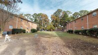 Located at 1840 Carriage Ln in Charleston, South Carolina, this single-story brick property sits within a well-established neighborhood in the 29407 zip code. The exterior showcases classic red brick construction, lending the buildings a timeless, durable aesthetic characteristic of the region.

A defining feature of this property is the generous open courtyard situated between the two parallel brick structures. The shared outdoor space offers ample room for recreation and relaxation, bordered by neatly trimmed shrubbery along the building foundations. A flagpole stands as a central landmark within the courtyard, while a chain-link fence defines the rear perimeter.

The grounds are framed by a striking canopy of mature trees, including tall pines and deciduous hardwoods displaying seasonal autumn foliage in warm golden and orange hues. This natural backdrop provides both shade and a sense of privacy, creating a tranquil atmosphere. The surrounding greenery contributes to the overall peaceful ambiance typical of this part of Charleston.

Each building features evenly spaced windows that allow for natural light throughout the interior units, and HVAC equipment is visible along the building exteriors. The property's layout, mature landscaping, and classic brick design come together to create a functional and quietly charming residential environment in West Ashley, Charleston.