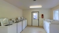 This single-floor laundry facility is designed with functionality and practicality in mind. The room features a row of top-load washing machines aligned along one wall, complemented by dryers positioned on the opposite side, providing ample capacity for multiple users. The space is finished with light-colored walls and durable flooring, creating a clean and well-maintained environment. Bright fluorescent overhead lighting ensures the room is well-lit throughout. A windowed exterior door allows natural light to filter in while providing ventilation and a view of the surrounding outdoor area. Safety is addressed with a prominently mounted fire extinguisher near the entry point. The overall layout is open and efficiently organized, allowing for easy movement between machines. The neutral, minimalist aesthetic keeps the focus on utility, making this a straightforward and accessible shared laundry space suited for communal use.
