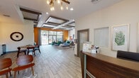 This single-story community clubhouse and leasing office is designed with a contemporary aesthetic that blends modern architecture with vibrant, mid-century-inspired furnishings. The space is defined by its striking coffered ceiling system, featuring layered geometric frames with dark trim accents, complemented by statement pendant chandeliers with exposed Edison-style bulbs that add warmth and visual interest.

Wood-look tile flooring runs throughout the open-plan interior, lending a cohesive and easy-to-maintain surface underfoot. Bold orange accent walls provide energetic contrast against the crisp white walls and neutral tones that dominate much of the space. The main lounge area is generously proportioned, furnished with tufted ottomans, a plush sofa with patterned throw pillows, and rich emerald-green velvet chairs that anchor the seating arrangement near floor-to-ceiling windows.

These expansive glazed panels and sliding doors frame direct views of the outdoor pool and patio, flooding the interior with natural light. A dedicated dining nook with a round table and sculptural seating sits adjacent to a kitchenette area, while a secondary lounge features an orange loveseat and artwork. The reception desk area offers a welcoming entry point, and additional rooms provide private or semi-private spaces for meetings or office use.
