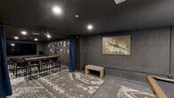 Located at 525 Main St in Hartford, CT, this single-level entertainment space is a thoughtfully designed, cinema-inspired lounge that blends screening room functionality with social leisure amenities. The interior is unified by a dramatic dark color palette — charcoal walls, black ceilings, and concrete accents — creating an intimate, immersive atmosphere throughout.

The screening area is the centerpiece of the space, equipped with a large flat-panel display and flanked by rows of mesh-back seating arranged for optimal viewing. The surrounding walls are lined with framed movie posters, reinforcing the cinematic theme. Recessed lighting provides controlled illumination without disrupting the theater ambiance.

A blue drape curtain serves as a flexible divider, separating the screening zone from a more casual lounge and social area. This secondary zone features a pool table positioned atop a bold geometric area rug, a built-in bar counter with bar-height seating, and additional wall-mounted artwork including a large abstract canvas that adds a vibrant contrast to the otherwise muted tones.

Practical touches include clearly marked exits, HVAC venting, and ample open floor space for circulation. The combination of entertainment-focused amenities, art-forward decor, and versatile layout makes this space well-suited for private screenings, social gatherings, or recreational use.