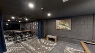 Located at 525 Main St in Hartford, CT, this single-level entertainment space is a thoughtfully designed, cinema-inspired lounge that blends screening room functionality with social leisure amenities. The interior is unified by a dramatic dark color palette — charcoal walls, black ceilings, and concrete accents — creating an intimate, immersive atmosphere throughout.

The screening area is the centerpiece of the space, equipped with a large flat-panel display and flanked by rows of mesh-back seating arranged for optimal viewing. The surrounding walls are lined with framed movie posters, reinforcing the cinematic theme. Recessed lighting provides controlled illumination without disrupting the theater ambiance.

A blue drape curtain serves as a flexible divider, separating the screening zone from a more casual lounge and social area. This secondary zone features a pool table positioned atop a bold geometric area rug, a built-in bar counter with bar-height seating, and additional wall-mounted artwork including a large abstract canvas that adds a vibrant contrast to the otherwise muted tones.

Practical touches include clearly marked exits, HVAC venting, and ample open floor space for circulation. The combination of entertainment-focused amenities, art-forward decor, and versatile layout makes this space well-suited for private screenings, social gatherings, or recreational use.