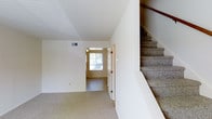 This two-story home offers a straightforward and functional layout designed for comfortable living. The main level features an open living area finished with wall-to-wall carpeting in neutral tones, creating a clean and cohesive feel throughout the space. Smooth white walls and a flat ceiling with a visible HVAC vent contribute to the bright, airy atmosphere of the interior.

Toward the front of the home, a welcoming entryway benefits from a window that allows natural light to filter into the space, illuminating the transition area between the main living zone and the front door. A white interior door near the entryway adds a classic, clean detail to the overall design.

A carpeted staircase with a warm wood handrail runs along one side of the main level, providing access to the second floor. The staircase is neatly integrated into the layout, making efficient use of the available space without disrupting the flow of the main level. Overall, the home presents a neutral palette and practical design that provides a versatile foundation for a variety of living arrangements.