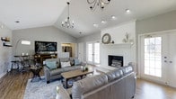 This single-story home showcases a thoughtfully designed open-concept layout that balances farmhouse character with contemporary living. The living room serves as the heart of the space, anchored by a striking whitewashed brick fireplace with a crisp white mantel, adding a cozy yet refined focal point to the room. Vaulted ceilings rise dramatically overhead, creating an expansive, airy atmosphere, while two elegant black chandelier-style light fixtures provide warm, layered illumination alongside recessed lighting. Large French doors bathe the space in abundant natural light and offer a seamless connection to the outdoors. The living area is generously proportioned, comfortably accommodating two large leather sofas arranged around a rustic wood-and-metal coffee table, all set atop a patterned area rug. Soft gray walls complement the neutral tones throughout, lending the space a calm, welcoming ambiance. A glimpse into the kitchen reveals modern stainless steel appliances, and a charming chalkboard sign adds a personalized, homey touch near the entryway. The overall design strikes a harmonious balance between rustic farmhouse charm and clean, modern finishes, making this a warm and inviting home.