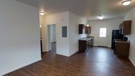 This single-story space presents an open-concept layout where the kitchen and living area flow seamlessly together. The flooring throughout is a rich, dark hardwood-style material that adds warmth and contrast to the neutral white walls and ceilings.

The kitchen is equipped with dark espresso-toned cabinetry offering generous storage, complemented by a matching dark countertop. A pendant light hangs above, adding a functional focal point to the space. The kitchen also features a sink positioned beneath a window, allowing natural light to illuminate the work area. A range and additional upper and lower cabinetry line the walls, maximizing storage and functionality.

An electrical panel is visibly mounted near the entrance, a practical and accessible feature of the layout. A white entry door at the far end of the kitchen provides additional natural light and access.

The open living area benefits from a second window fitted with white blinds, ensuring balanced light distribution across the space. The combination of dark flooring, dark cabinetry, and bright walls creates a clean, functional aesthetic throughout this straightforward, single-level layout.