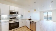 This thoughtfully designed single-floor unit offers a clean, contemporary aesthetic throughout. The kitchen serves as a standout centerpiece, featuring a full suite of stainless steel appliances — including a refrigerator, range, microwave, and dishwasher — complemented by crisp white cabinetry that extends to the ceiling for ample storage. A granite-topped peninsula with an undermount sink provides both workspace and a casual dining option, while globe pendant lights in a warm gold finish add a stylish touch overhead. A soft blue-gray subway tile backsplash ties the kitchen together with subtle elegance. Light hardwood-style flooring flows through the main living and kitchen areas, giving the space a bright, airy feel. The living area opens to a private balcony through full-height sliding glass doors, offering outdoor views and natural light. Bedrooms are finished with plush carpet flooring and include generous closet space with built-in shelving and hanging rods. The bathroom is functional and well-appointed, featuring a walk-in shower, a granite-topped vanity, and additional built-in shelving for convenient storage. Recessed lighting throughout ensures consistent, even illumination across all areas of the unit.
